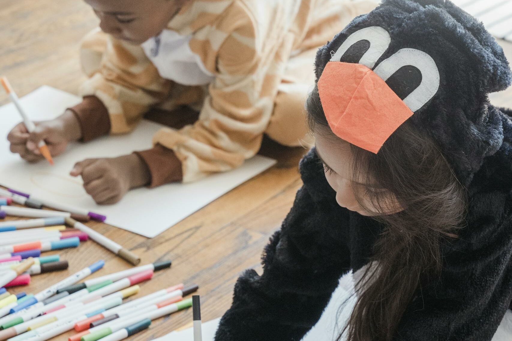 Two kids in animal costumes drawing colorful pictures on white paper indoors.